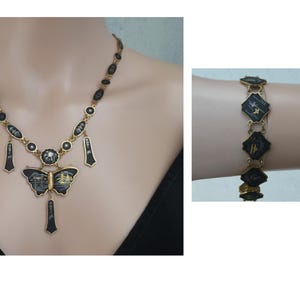 Op de afbeelding: Een gouden ketting met een zwarte vlinder hanger en zwarte en gouden accenten. De ketting is om de nek van een model. Een bijpassende armband met zwarte en gouden accenten is om de pols van een model.