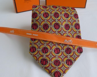 Könnte beinhalten: Eine Seidenkrawatte mit einem sich wiederholenden geometrischen Muster aus roten kreisförmigen Formen und gelben Halbmondmotiven auf taupefarbenem Grund. Ein orangefarbenes Band mit dem Text HERMES-PARIS und einem Kutschenlogo liegt quer.