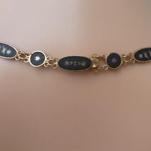 Op de afbeelding: Een gouden ketting met zwarte ovale hangers. Elke hanger heeft een wit ontwerp en gouden letters. De sluiting is een gouden ovaal met gouden letters.