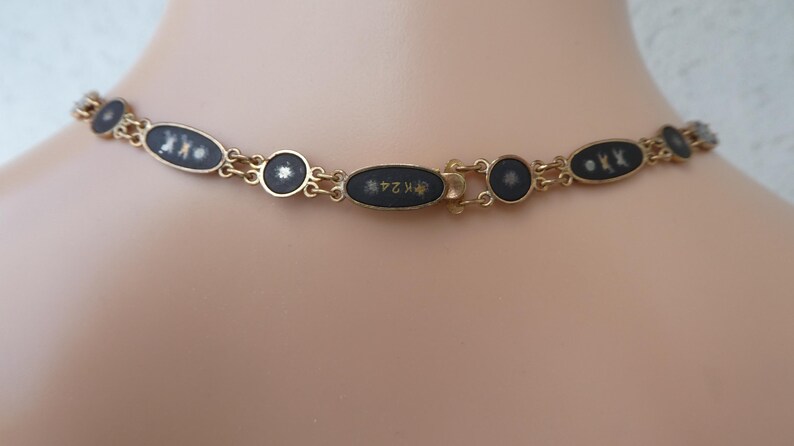 Op de afbeelding: Een gouden ketting met zwarte ovale hangers. Elke hanger heeft een wit ontwerp en gouden letters. De sluiting is een gouden ovaal met gouden letters.