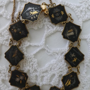 Op de afbeelding: Een gouden kettingarmband met zwarte emaille bedels met Japanse sc&egrave;nes in goud. De bedels zijn verbonden door gouden schakels. De armband heeft een sluiting met de markering "K 24".