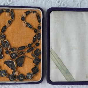 Op de afbeelding: Een zwarte en gouden ketting met een vlinderhanger in een paarse fluwelen doos. De ketting is gemaakt van kleine, ronde zwarte kralen en gouden accenten. De vlinderhanger is gemaakt van zwart emaille en goud.