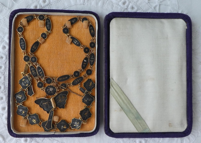 Op de afbeelding: Een zwarte en gouden ketting met een vlinderhanger in een paarse fluwelen doos. De ketting is gemaakt van kleine, ronde zwarte kralen en gouden accenten. De vlinderhanger is gemaakt van zwart emaille en goud.