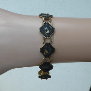 Op de afbeelding: Een gouden armband met zwarte en witte emaille vierkanten met een vogelontwerp.