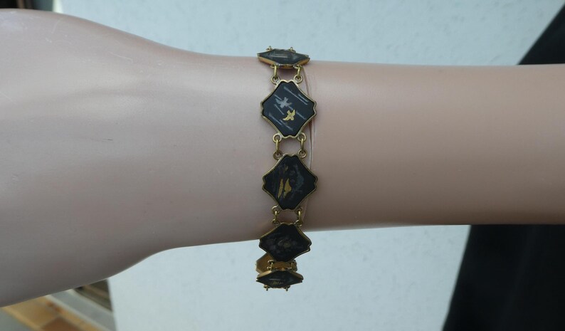Op de afbeelding: Een gouden armband met zwarte en witte emaille vierkanten met een vogelontwerp.