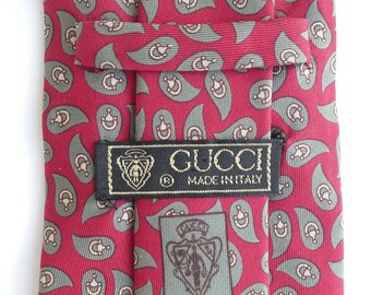 May include: Red silk tie with a paisley pattern in green and beige. The tie has a black label with the text "GUCCI MADE IN ITALY".