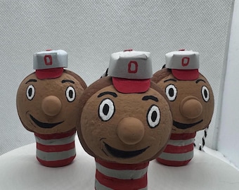 May include: Three handmade figurines of a mascot with a brown face, a red and white striped body, and a hat with a red brim and a gray top with a red "O". The figurines have a smiling face and a round nose.