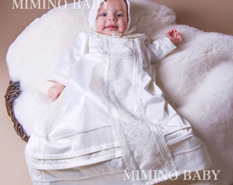 May include: A white satin baby christening gown with intricate lace detailing down the front and on the matching bonnet. The gown has long sleeves and is trimmed with delicate stitching. The text "MIMINO BABY" is visible.