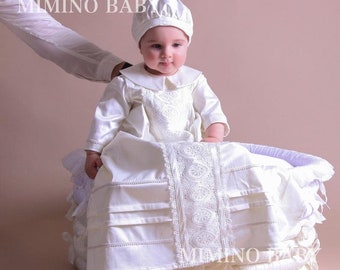 Può includere: Un abito da battesimo in raso bianco con bordino in pizzo e un berretto abbinato. L'abito ha una gonna a pieghe e un piccolo colletto. Il bambino è seduto in un cesto di vimini bianco.
