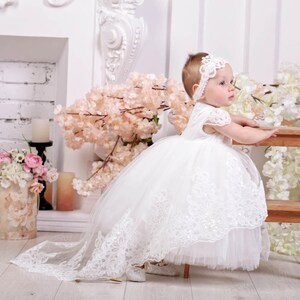 May include: A white tulle and lace baby dress with a long train. The dress has a white lace bodice and a white tulle skirt. The baby is wearing a white headband with a flower design.
