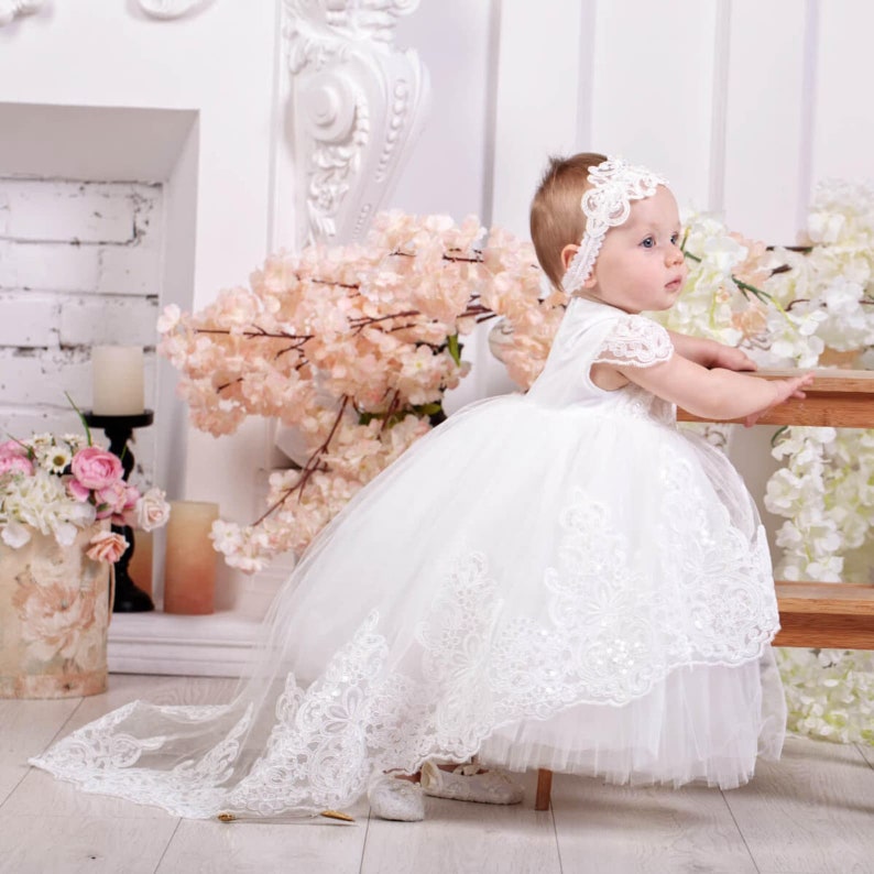 May include: A white tulle and lace baby dress with a long train. The dress has a white lace bodice and a white tulle skirt. The baby is wearing a white headband with a flower design.