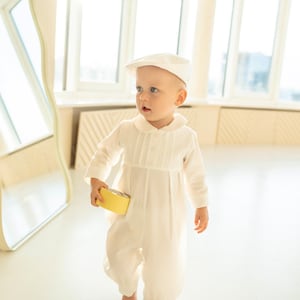 May include: A baby wearing a white long-sleeved jumpsuit with a white hat and holding a yellow toy.