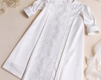 May include: A white satin christening gown with intricate lace detailing down the front. The gown features long sleeves with lace cuffs and a pleated skirt. A matching lace bonnet is also visible.