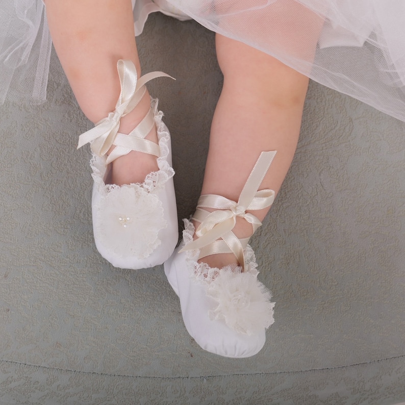 May include: A pair of white baby shoes with lace trim and satin ribbons. The shoes have a heart-shaped embellishment on the toe.