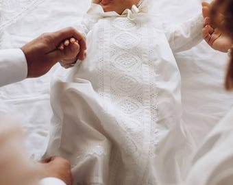 May include: A baby wearing a white christening gown with lace trim, lying on a white blanket. The baby is being held by two adults, one on each side.