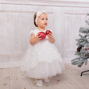 May include: A young girl in a white dress with lace trim holds two red Christmas baubles. She is wearing a white headband with a flower on it. The girl is standing in front of a white wall with a Christmas tree in the background.