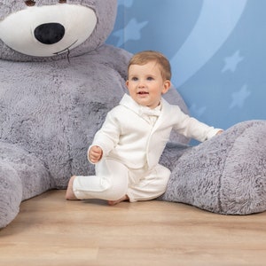 May include: A baby boy wearing a white suit sits next to a large grey teddy bear. The baby is smiling and looking to the side. The teddy bear is sitting on a wooden floor.
