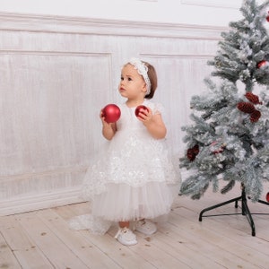 May include: A young girl wearing a white dress with lace and tulle, holding two red Christmas baubles. She is standing in front of a snowy Christmas tree.