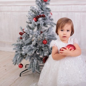 May include: A young girl in a white dress with lace trim holds two red Christmas baubles in front of a snowy Christmas tree.