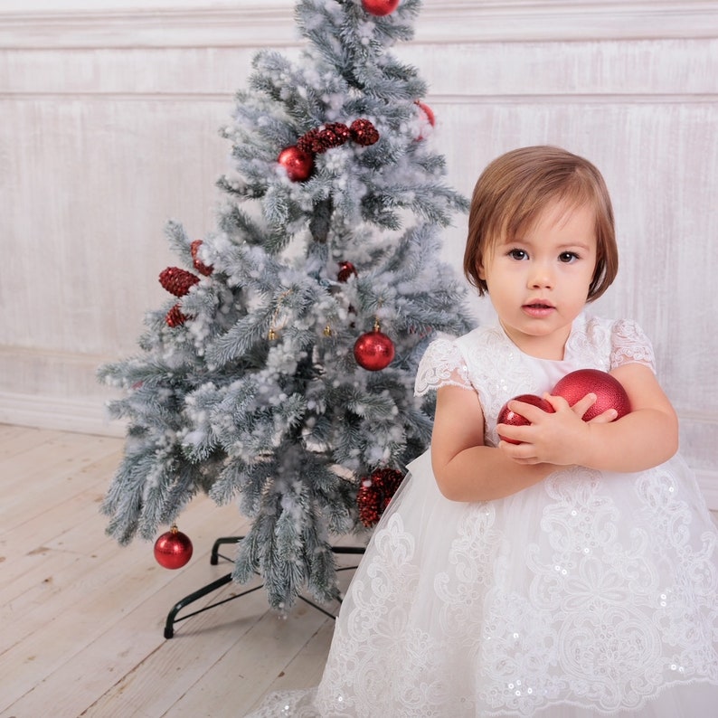 May include: A young girl in a white dress with lace trim holds two red Christmas baubles in front of a snowy Christmas tree.