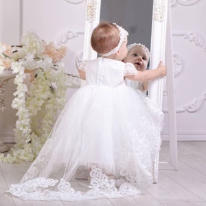 May include: A young girl in a white tulle dress with lace trim and a long train looks at her reflection in a mirror.