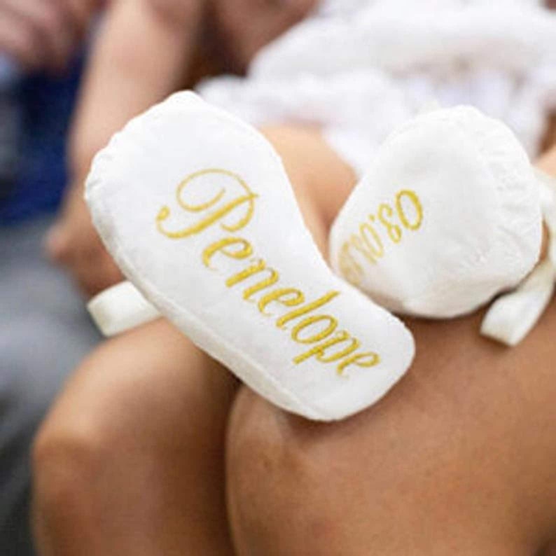May include: White baby booties with gold embroidery. The booties are personalised with the name "Penelope" on one bootie and "No. 1" on the other.