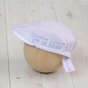 May include: A white newsboy cap with light blue embroidered text reading "Cristian Lorenzo 07.15.2023." The cap is displayed on a wooden form. A small tag with text is attached to the cap.