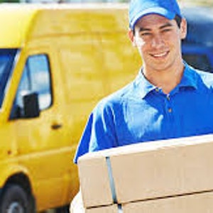 May include: A delivery driver wearing a blue uniform and cap is smiling and holding a stack of brown cardboard boxes. He is standing in front of a yellow delivery van.