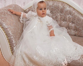 Op de afbeelding: Een witte doopjurk met kantdetails en een bijpassende muts. De jurk heeft een volle rok en is versierd met pailletten. De jurk wordt gepresenteerd op een witte en gouden chaise longue.