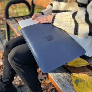 May include: A dark blue leather laptop sleeve with an Apple logo. The sleeve is closed and being held by a person sitting on a wooden bench.