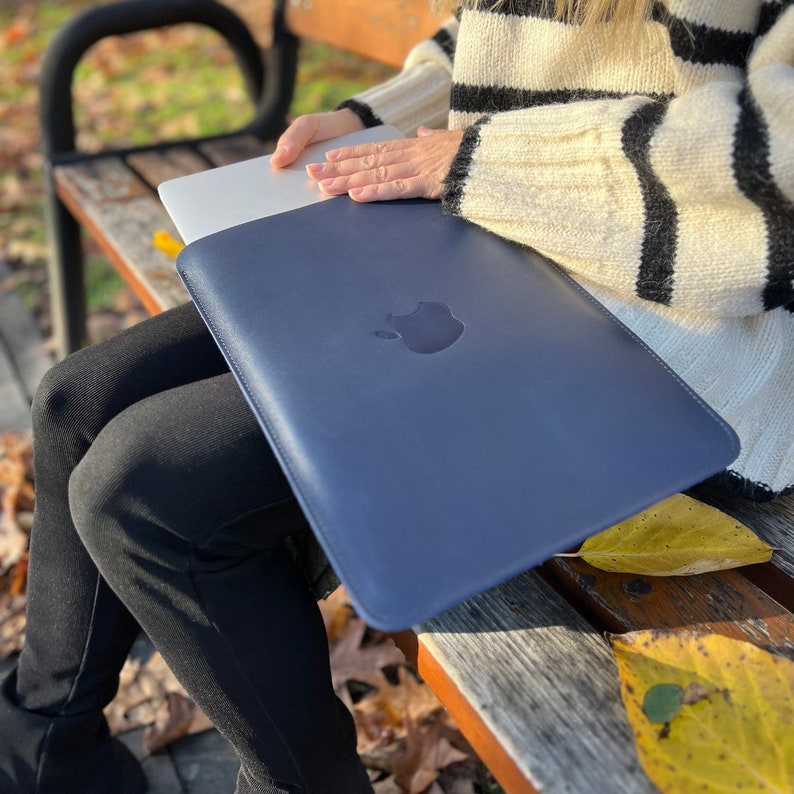 May include: A dark blue leather laptop sleeve with an Apple logo. The sleeve is closed and being held by a person sitting on a wooden bench.