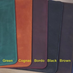 K&ouml;nnte beinhalten: Eine Auswahl an Schreibtischunterlagen aus Leder in verschiedenen Farben: Gr&uuml;n, Cognac, Bordeaux, Schwarz, Braun und Blau. Jede Unterlage ist rechteckig mit abgerundeten Ecken und gen&auml;hten Kanten. Die Farbbezeichnungen sind in gelber Schrift dargestellt.