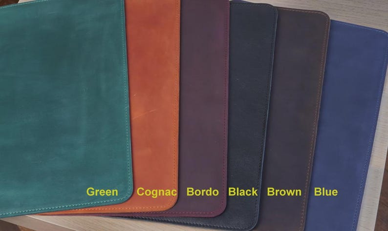 K&ouml;nnte beinhalten: Eine Auswahl an Schreibtischunterlagen aus Leder in verschiedenen Farben: Gr&uuml;n, Cognac, Bordeaux, Schwarz, Braun und Blau. Jede Unterlage ist rechteckig mit abgerundeten Ecken und gen&auml;hten Kanten. Die Farbbezeichnungen sind in gelber Schrift dargestellt.