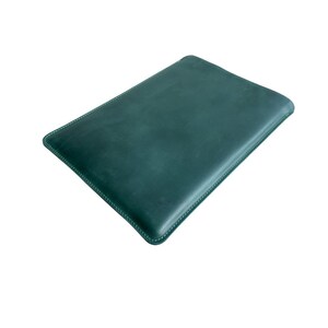 K&ouml;nnte beinhalten: Eine dunkelgr&uuml;ne Lederh&uuml;lle mit wei&szlig;en N&auml;hten an den Kanten. Die rechteckige H&uuml;lle ist f&uuml;r ein Tablet oder ein &auml;hnliches Ger&auml;t konzipiert. Das Leder hat eine leicht strukturierte Oberfl&auml;che.