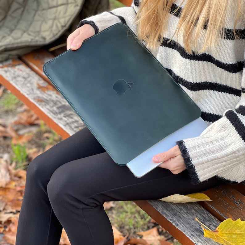 K&ouml;nnte beinhalten: Eine Person h&auml;lt eine dunkelgr&uuml;ne Lederh&uuml;lle f&uuml;r einen Laptop mit einem Apple-Logo. Die H&uuml;lle liegt auf einem silbernen Laptop.