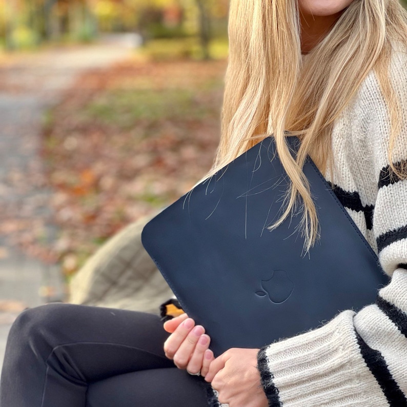 K&ouml;nnte beinhalten: Eine Frau h&auml;lt eine dunkelblaue Laptoptasche mit einem Apple-Logo. Die Tasche ist aus weichem, strapazierf&auml;higem Material gefertigt und eignet sich perfekt zum Schutz Ihres Laptops.