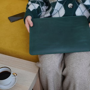 K&ouml;nnte beinhalten: Eine gr&uuml;ne Ledertasche f&uuml;r Laptops mit einem dezenten gepr&auml;gten Logo. Die Tasche ist geschlossen und wird von einer Person gehalten, die einen wei&szlig;en und gr&uuml;nen Pullover tr&auml;gt.