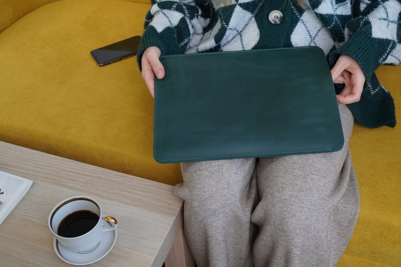 K&ouml;nnte beinhalten: Eine gr&uuml;ne Ledertasche f&uuml;r Laptops mit einem dezenten gepr&auml;gten Logo. Die Tasche ist geschlossen und wird von einer Person gehalten, die einen wei&szlig;en und gr&uuml;nen Pullover tr&auml;gt.