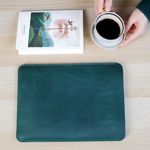 K&ouml;nnte beinhalten: Eine teal-farbene Laptop-H&uuml;lle aus Leder auf einer hellen Holzoberfl&auml;che. Ein Stapel B&uuml;cher mit einer floralen Illustration und dem Text "RIVER BOOKS" befindet sich neben der H&uuml;lle. Eine Tasse Kaffee auf einer Untertasse wird im Hintergrund gehalten.