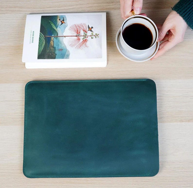 K&ouml;nnte beinhalten: Eine teal-farbene Laptop-H&uuml;lle aus Leder auf einer hellen Holzoberfl&auml;che. Ein Stapel B&uuml;cher mit einer floralen Illustration und dem Text "RIVER BOOKS" befindet sich neben der H&uuml;lle. Eine Tasse Kaffee auf einer Untertasse wird im Hintergrund gehalten.