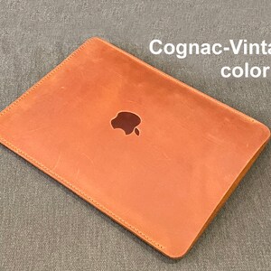 May include: A brown leather laptop sleeve with a stitched edge and the Apple logo in the center. The sleeve is labeled "Cognac-Vintage color".
