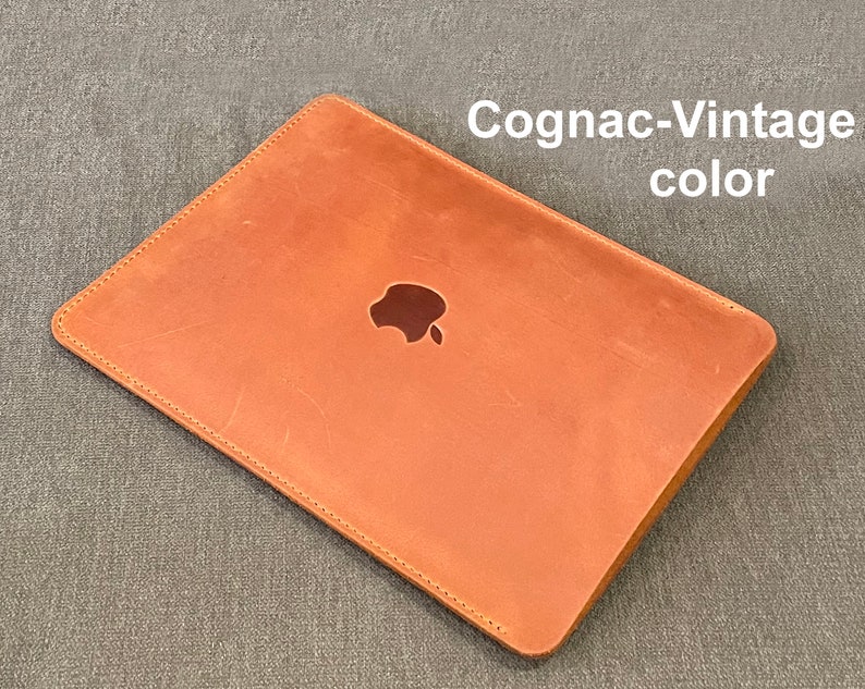 May include: A brown leather laptop sleeve with a stitched edge and the Apple logo in the center. The sleeve is labeled "Cognac-Vintage color".
