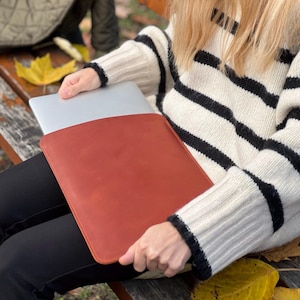 K&ouml;nnte beinhalten: Eine rotbraune Laptop-H&uuml;lle aus Leder. Die H&uuml;lle wird von einer Person gehalten, die einen cremefarbenen Pullover mit schwarzen horizontalen Streifen tr&auml;gt. Der Laptop ist teilweise in der H&uuml;lle sichtbar. Der Hintergrund umfasst eine Holzbank und Herbstbl&auml;tter.