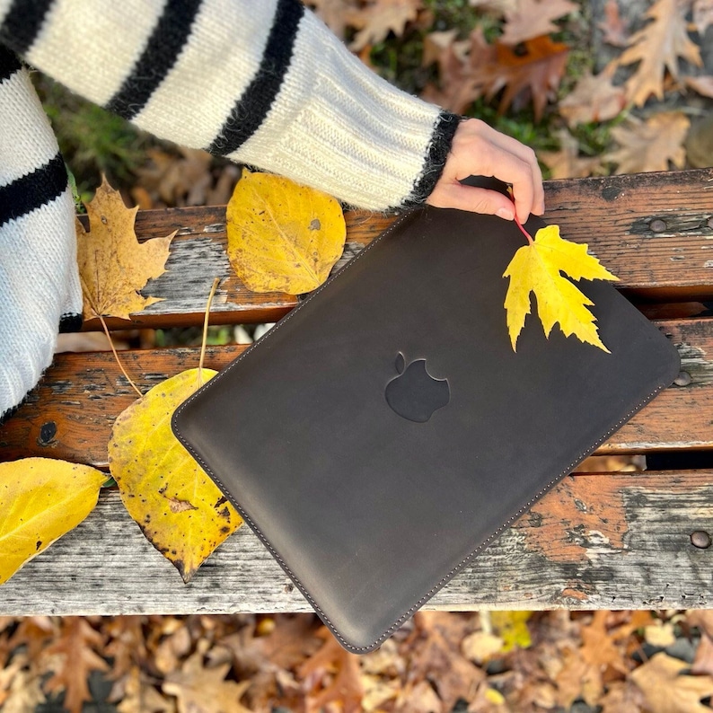 K&ouml;nnte beinhalten: Eine braune Lederh&uuml;lle f&uuml;r Laptops mit einem gen&auml;hten Rand und einem Apple-Logo. Die H&uuml;lle liegt auf einer Holzbank mit verstreuten gelben Bl&auml;ttern.