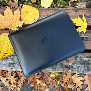 May include: Black leather laptop sleeve with an Apple logo. The sleeve is lying on a wooden surface with autumn leaves scattered around it.