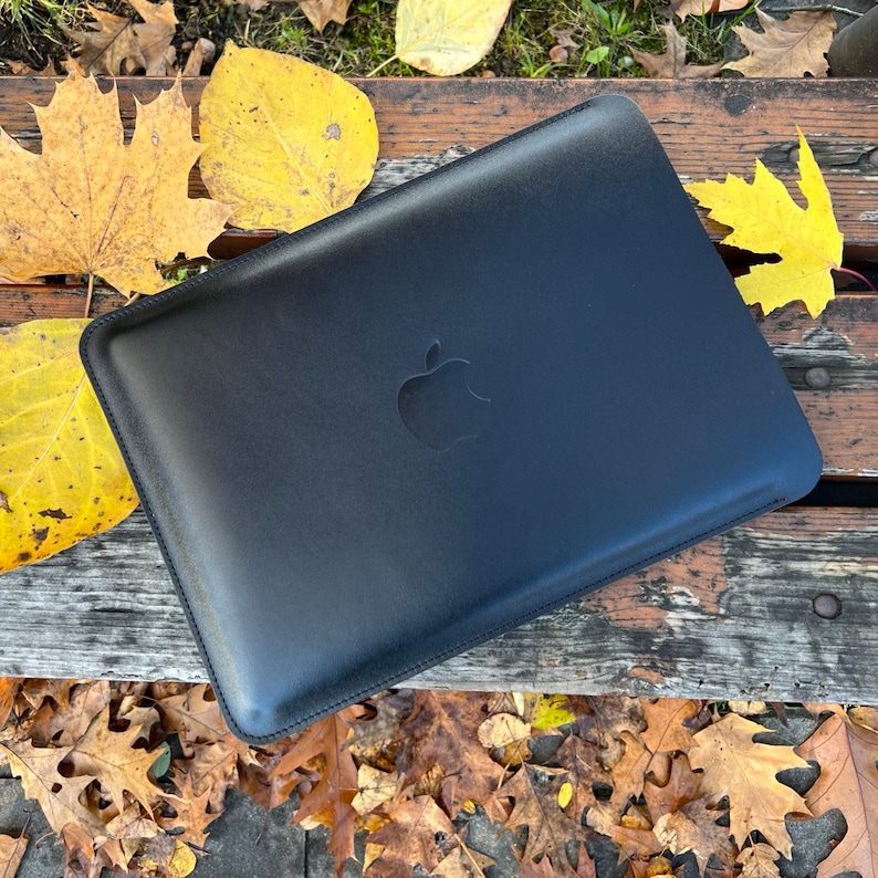 May include: Black leather laptop sleeve with an Apple logo. The sleeve is lying on a wooden surface with autumn leaves scattered around it.