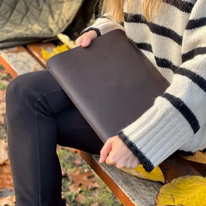 K&ouml;nnte beinhalten: Eine dunkelbraune Laptop-H&uuml;lle aus Leder mit einer gen&auml;hten Bord&uuml;re. Die H&uuml;lle wird von einer Person gehalten, die einen schwarz-wei&szlig; gestreiften Pullover tr&auml;gt. Der Hintergrund umfasst eine Holzbank und gefallene gelbe Bl&auml;tter.
