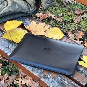 May include: A black leather laptop sleeve with the Apple logo on the front. The sleeve is sitting on a wooden bench with fallen leaves around it.