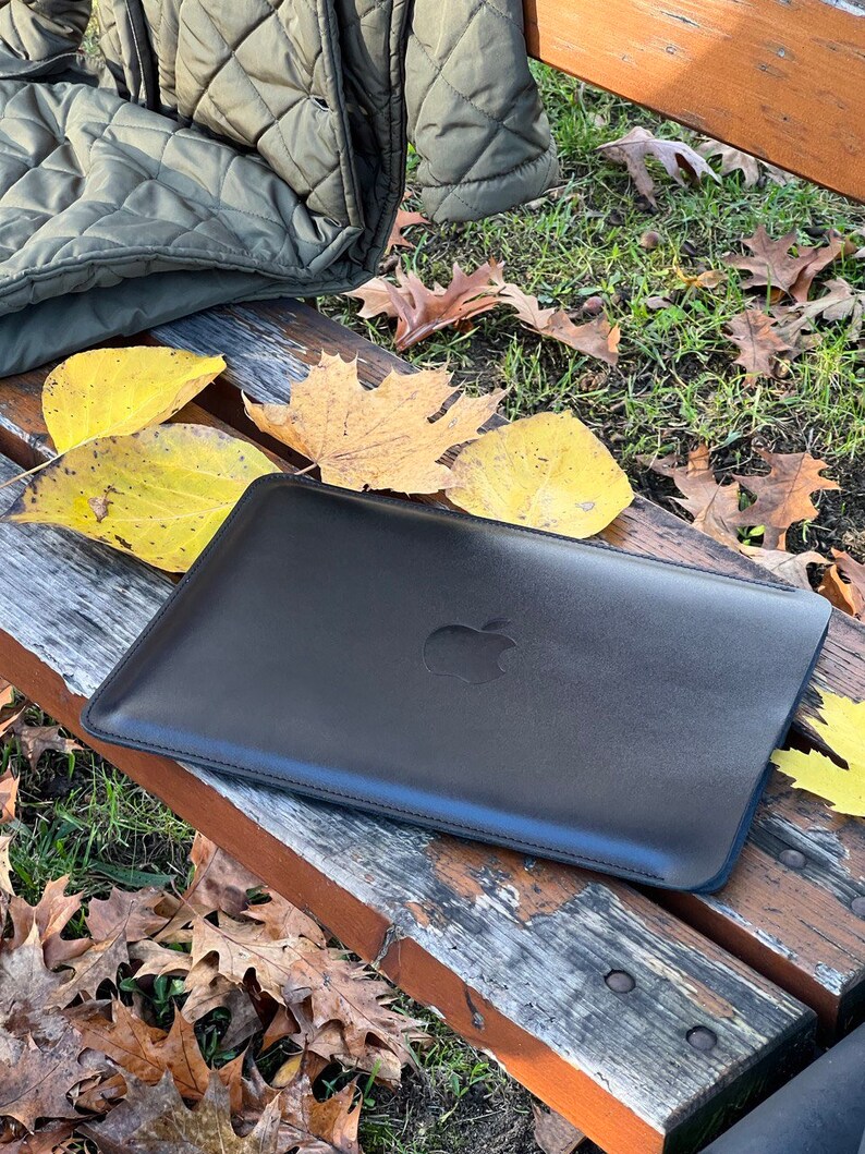 May include: A black leather laptop sleeve with the Apple logo on the front. The sleeve is sitting on a wooden bench with fallen leaves around it.