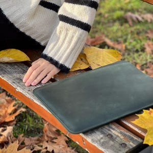 K&ouml;nnte beinhalten: Eine dunkelgr&uuml;ne Laptop-H&uuml;lle aus Leder liegt auf einer Holzbank, umgeben von gelben Herbstbl&auml;ttern. Die rechteckige H&uuml;lle hat abgerundete Ecken und eine glatte Oberfl&auml;che. Eine Person tr&auml;gt einen cremefarbenen Pullover mit schwarzen Streifen.